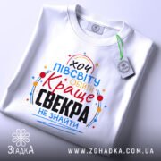 Футболка для найкращого свекра, білого кольору, зображена з верхнього ракурсу, на ній є етикетка, фон світло-сірий, принт яскравий, з текстом "хочу півсвіту обійти Краще свекра не знайти".