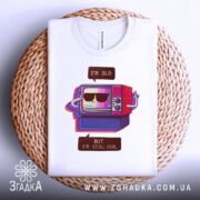 Футболка для олдів, білого кольору, зображена зверху на плетеній основі, фон світлий, принт з телевізором у ретро стилі, напис 'I'M OLD' та 'BUT I'M STILL COOL'.