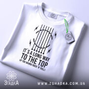 Футболка для рокера, біла, складена на поверхні, фон світлий, принт чорного кольору з текстом "IT'S A LONG WAY TO THE TOP (IF YOU WANNA ROCK 'N' ROLL)" та графічними елементами, на футболці підвісна етикетка.