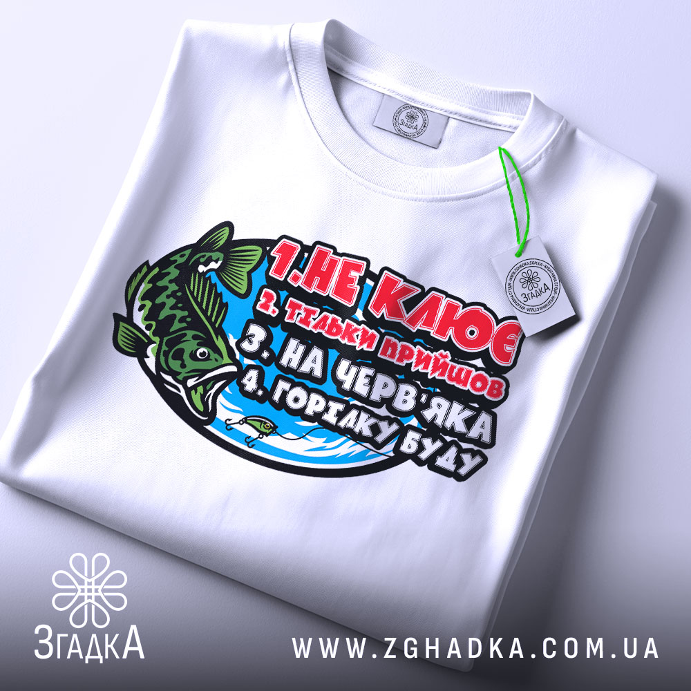 Футболка для риболовлі, білого кольору, зображена зверху на столі, фон нейтральний, принт з рибою та текстом, на футболці є етикетка.