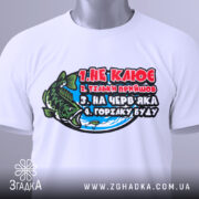 Футболка для риболовлі, білого кольору, зображена зверху на столі, фон нейтральний, принт з рибою та текстом, на футболці є етикетка.