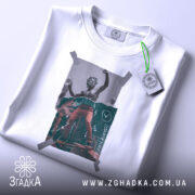 Футболка для скейтерів, біла, складена на поверхні, фон світло-сірий, принт з малюнком скейтера в червоних штанах, обклеєний скотчем, з етикеткою зеленого кольору.