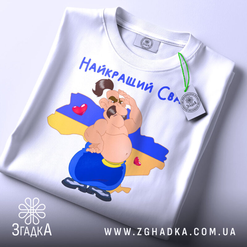 Футболка для свата, біла, складена на поверхні, фон нейтральний, принт з веселим персонажем на фоні карти України, текст 'Найкращий Сват' зверху, етикетка на рукаві.