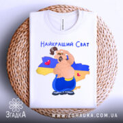 Футболка для свата, біла, складена на плетеній основі, фон нейтральний, принт з веселим персонажем на фоні карти України, текст 'Найкращий Сват' зверху, деталі принта добре проглядаються.