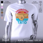 Футболка для тата білого кольору, зображена з близького ракурсу, на манекені. Принт містить зображення двох кулаків та текст 'PROMOTED TO TATO' у яскравих кольорах на фоні з градієнтом.