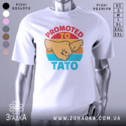 Футболка для тата білого кольору, зображена з близького ракурсу, на манекені. Принт містить зображення двох кулаків та текст 'PROMOTED TO TATO' у яскравих кольорах на фоні з градієнтом.