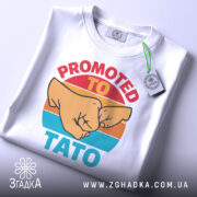 Футболка для тата білого кольору, складена на поверхні, з видимим етикеткою. Принт з двома кулаками та текстом 'PROMOTED TO TATO' яскраво виділяється на фоні.