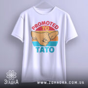 Футболка для тата білого кольору, підвішена на вішалці, з фронтального ракурсу. Принт з двома кулаками та текстом 'PROMOTED TO TATO' чітко видно на фоні.