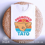 Футболка для тата білого кольору, складена на плетеній кошику. Принт з двома кулаками та текстом 'PROMOTED TO TATO' яскраво контрастує з фоном.