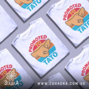 Футболка для тата білого кольору, зображена з близького ракурсу, на манекені. Принт містить зображення двох кулаків та текст 'PROMOTED TO TATO' у яскравих кольорах на фоні з градієнтом.