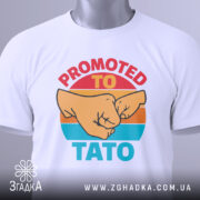 Футболка для тата білого кольору, складена на поверхні, з видимим етикеткою. Принт з двома кулаками та текстом 'PROMOTED TO TATO' яскраво виділяється на фоні.