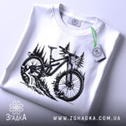 Футболка для велосипедиста, білого кольору, складена на поверхні, фон світлий, принт з чорним малюнком велосипеда на фоні лісу, з етикеткою зеленого кольору.