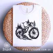 Футболка для велосипедиста, білого кольору, складена на плетеній кошику, фон світлий, принт з чорним малюнком велосипеда на фоні лісу.
