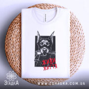 Футболка доберман живи життя, біла футболка складена на плетеній кошику, зображення добермана з пістолетом у пащі на чорному фоні, текст 'ЖИВИ ЖИТТЯ' червоним кольором.