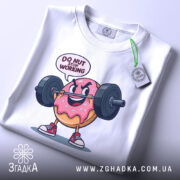 Футболка донат спорт мотивація, складена біла футболка з принтом веселого пончика, що піднімає штангу, текст 'DO NUT STOP WORKING' над принтом, на фоні світло-сірого кольору, з етикеткою зеленого кольору.