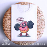 Футболка донат спорт мотивація, складена біла футболка з принтом веселого пончика, що піднімає штангу, текст 'DO NUT STOP WORKING' над принтом, на фоні плетеного кошика, що має світло-сірий колір.