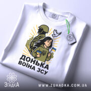 Футболка донька воїна ЗСУ, білого кольору, зображена зверху, фон світло-сірий, принт з дівчиною, солдатом та голубом, на футболці є етикетка з зеленим шнурком.