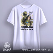 Футболка донька воїна ЗСУ, білого кольору, підвішена на вішалці, фон світло-сірий, принт з дівчиною, солдатом та голубом, яскраві жовті промені навколо.