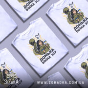 Футболка донька воїна ЗСУ, білого кольору, зображена на манекені, фон світло-сірий, принт з дівчиною, солдатом та голубом, яскраві жовті промені навколо.