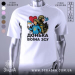 Футболка донька воїна ЗСУ, біла, зображена з фронтального ракурсу на манекені, фон світло-сірий, принт з дівчиною у блакитному худі, яка тримає прапор України, поряд солдат у формі, на фоні силуети міста, червоне сердечко та голуб.