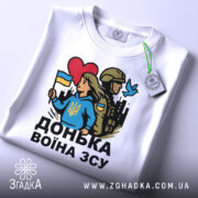 Футболка донька воїна ЗСУ, біла, зображена зверху, на ній видно етикетку з зеленим шнурком, фон світло-сірий, принт з дівчиною у блакитному худі, яка тримає прапор України, поряд солдат у формі, червоне сердечко та голуб.