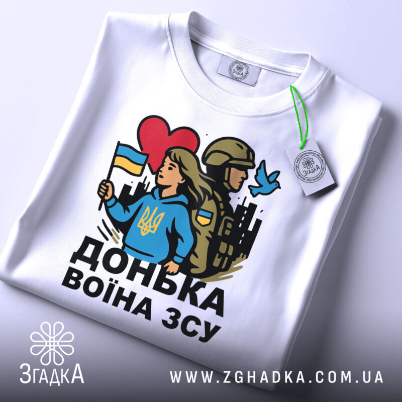 Футболка донька воїна ЗСУ, біла, зображена зверху, на ній видно етикетку з зеленим шнурком, фон світло-сірий, принт з дівчиною у блакитному худі, яка тримає прапор України, поряд солдат у формі, червоне сердечко та голуб.