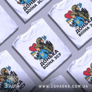 Футболка донька воїна ЗСУ, біла, зображена з фронтального ракурсу на манекені, фон світло-сірий, принт з дівчиною у блакитному худі, яка тримає прапор України, поряд солдат у формі, на фоні силуети міста, червоне сердечко та голуб.