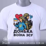 Футболка донька воїна ЗСУ, біла, зображена зверху, на ній видно етикетку з зеленим шнурком, фон світло-сірий, принт з дівчиною у блакитному худі, яка тримає прапор України, поряд солдат у формі, червоне сердечко та голуб.