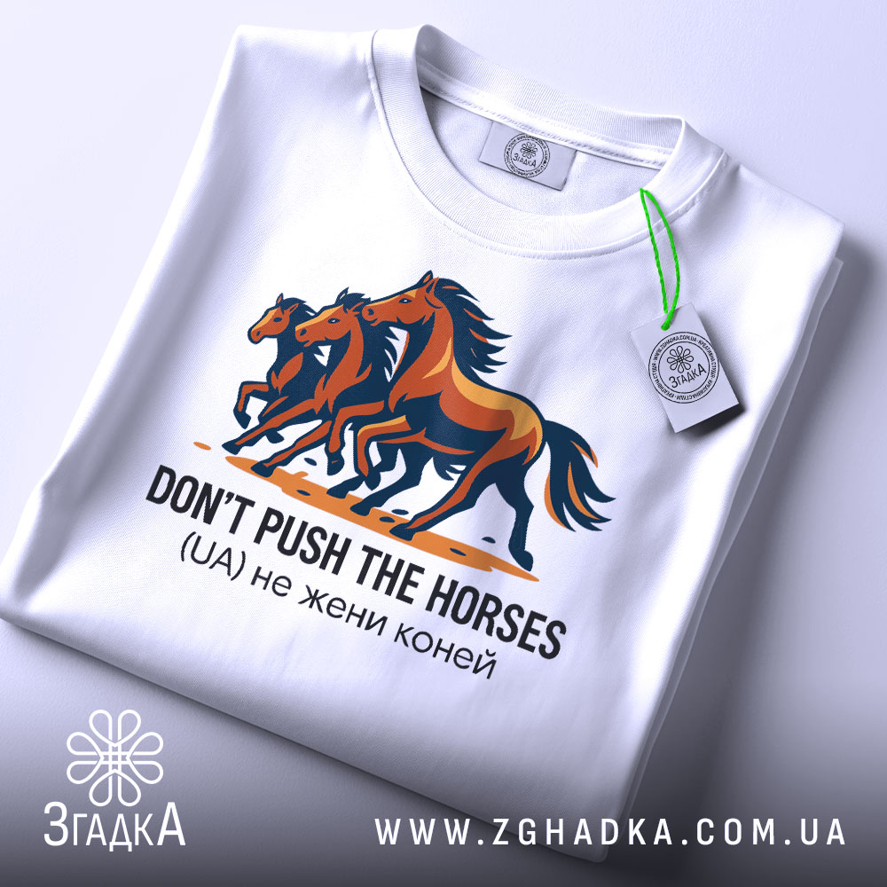 Футболка don't push the horses, білого кольору, зображена на столі, ракурс зверху, фон світло-сірий, принт з трьома конями, кольори принта - коричневий, чорний та оранжевий, текст під принтом чорного кольору, на футболці є етикетка зеленого кольору.