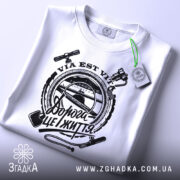 Футболка дорога це життя, складена біла футболка з чорним принтом, зображена на білому фоні, принт містить текст "Дорога це життя" та зображення інструментів, на футболці є етикетка з зеленим шнурком.