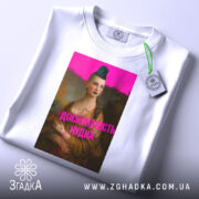 Футболка досконалість нудна, білого кольору, з верхнього ракурсу, на плоскій поверхні, фон світлий, принт з зображенням жінки з зеленим волоссям та написом 'ДОСКОНАЛІСТЬ НУДНА' яскраво-рожевого кольору, з етикеткою на комірці.