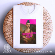 Футболка досконалість нудна, білого кольору, з верхнього ракурсу, на плоскій поверхні, фон нейтральний, принт з зображенням жінки з зеленим волоссям та написом 'ДОСКОНАЛІСТЬ НУДНА' яскраво-рожевого кольору, розташована на плетеній основі.