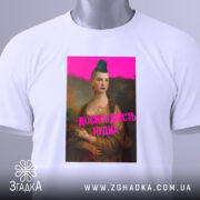 Футболка досконалість нудна, білого кольору, з верхнього ракурсу, на плоскій поверхні, фон світлий, принт з зображенням жінки з зеленим волоссям та написом 'ДОСКОНАЛІСТЬ НУДНА' яскраво-рожевого кольору, з етикеткою на комірці.