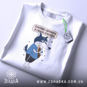 Футболка дружу з кукухою, біла, складена на поверхні, фон світло-сірий, принт з малюнком вовка у блакитній футболці, який тримає кукурудзу, текст над малюнком українською мовою, на футболці є етикетка з зеленим шнурком.