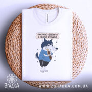 Футболка дружу з кукухою, біла, складена на плетеній кошику, фон білий, принт з малюнком вовка у блакитній футболці, який тримає кукурудзу, текст над малюнком українською мовою.