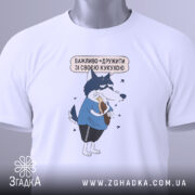 Футболка дружу з кукухою, біла, складена на поверхні, фон світло-сірий, принт з малюнком вовка у блакитній футболці, який тримає кукурудзу, текст над малюнком українською мовою, на футболці є етикетка з зеленим шнурком.