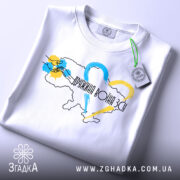 Футболка дружина воїна ЗСУ, білого кольору, складена на поверхні, фон світло-сірий, принт з контуром України, кольоровими серцями та написом, з етикеткою на комірці.