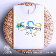 Футболка дружина воїна ЗСУ, білого кольору, складена на плетеній кошику, фон світло-сірий, принт з контуром України, кольоровими серцями та написом, видно комірець.