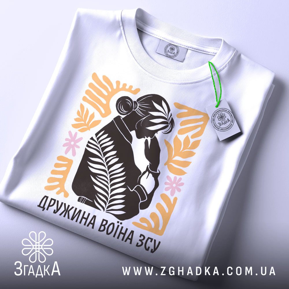 Футболка дружина воїна ЗСУ, біла, розкладена на поверхні, з видимим етикеткою, фон світло-сірий, принт з силуетом жінки, що тримає руку на обличчі, оточений рослинними елементами в жовтому та рожевому кольорах.