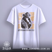 Футболка дружина воїна ЗСУ, біла, підвішена на вішалці, фон світло-сірий, принт з силуетом жінки, що тримає руку на обличчі, оточений рослинними елементами в жовтому та рожевому кольорах.