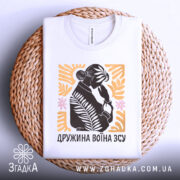 Футболка дружина воїна ЗСУ, біла, складена на плетеній кошику, фон світло-сірий, принт з силуетом жінки, що тримає руку на обличчі, оточений рослинними елементами в жовтому та рожевому кольорах.