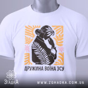 Футболка дружина воїна ЗСУ, біла, розкладена на поверхні, з видимим етикеткою, фон світло-сірий, принт з силуетом жінки, що тримає руку на обличчі, оточений рослинними елементами в жовтому та рожевому кольорах.