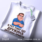Футболка дружина змусила прийти, біла, зображена зверху, на столі, фон світло-сірий, принт з малюнком сердитого чоловіка в синій футболці та текстом на українській мові, з етикеткою зеленого кольору.