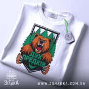 Футболка дух предків білого кольору, зображена зверху, на ній видно етикетку, фон світло-сірий, принт з ведмедем та лісом яскравий і детальний.
