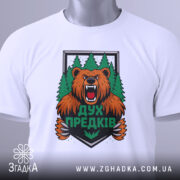 Футболка дух предків білого кольору, зображена зверху, на ній видно етикетку, фон світло-сірий, принт з ведмедем та лісом яскравий і детальний.