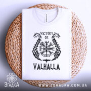 Футболка дух вікінга, біла, зображена зверху на плетеному кошику, фон білий, принт чорного кольору з написом 'VICTORY OR VALHALLA' та візерунками вікінгів.