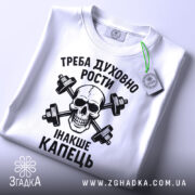 Футболка духовний ріст, білого кольору, складена на поверхні, фон світло-сірий, принт чорного кольору з черепом та гантелями, текст українською мовою, на етикетці зелена стрічка.
