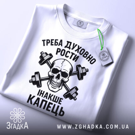 Футболка духовний ріст, білого кольору, складена на поверхні, фон світло-сірий, принт чорного кольору з черепом та гантелями, текст українською мовою, на етикетці зелена стрічка.