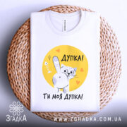 Футболка дупка ти моя, біла футболка, складена на плетеній кошику, принт з котом на жовтому фоні, текст "Дупка! Ти моя дупка!".