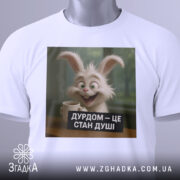 Футболка дурдом стан душі, біла, зображена на столі, фон світло-сірий, принт з усміхненим кроликом, що тримає чашку, текст на принті чорний з білими літерами, на комірці є етикетка.